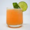 A vibrant orange Cantaloupe Basil Blaze cocktail in a glass, showcasing a blend of cantaloupe puree and mezcal, accented with a freshly smacked basil leaf and a lime wedge garnish, embodying a harmonious mix of sweet, smoky, and herbal notes.
