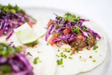 A vibrant image showing Cast-Iron Seared Flank Steak Tacos filled with juicy, caramelized flank steak, a colorful tangy red cabbage slaw and a creamy cheese sauce, all nestled in warm flour tortillas.