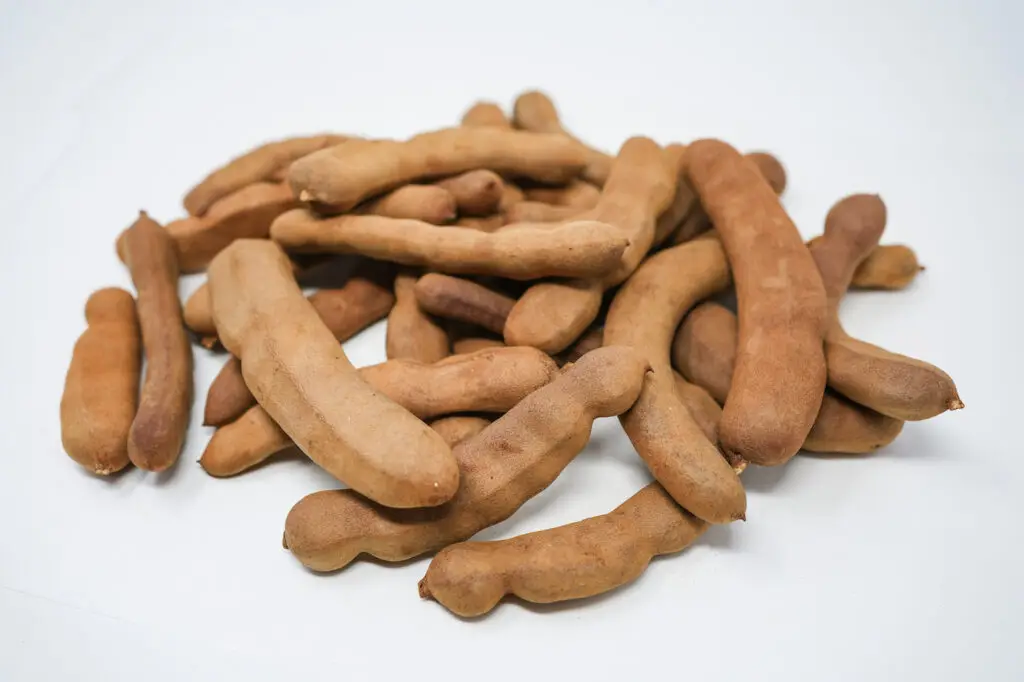 A close-up image of tamarind pods arranged neatly on a clean white background, showcasing their textured brown shells and curved, elongated shape.