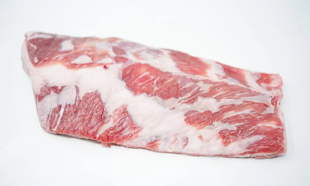 A raw, whole lamb breast displayed on a clean surface, showing its marbled fat and muscle with visible rib bones. The meat has a rich, pinkish-red color, indicative of its freshness and quality, ready for culinary preparation.