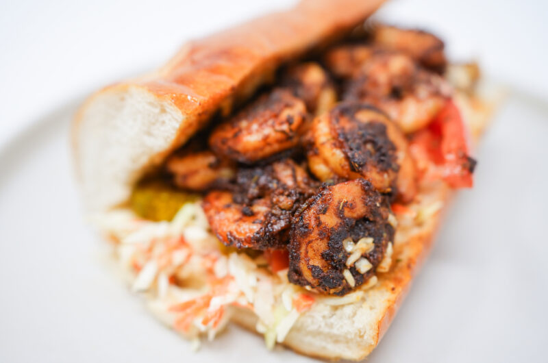 A Blackened Shrimp and Creamy Coleslaw Po'Boy sandwich with spicy shrimp, creamy coleslaw, bread and butter pickles, and fresh tomato slices in a ciabatta roll.