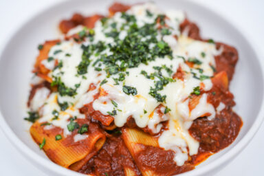 A bowl of Braised Beef Short Rib Rigatoni, featuring rigatoni pasta coated in a rich tomato sauce with tender shredded beef short ribs, garnished with melted mozzarella and fresh basil.