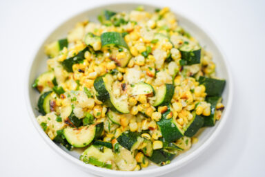 A vibrant salad featuring grilled corn kernels, zucchini strips, and cubes of asadero cheese, garnished with freshly chopped cilantro and green onions, all drizzled with a zesty lime vinaigrette on a white plate.