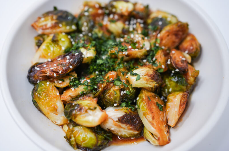 A plate of crispy Honey Sriracha Fried Brussels Sprouts, glistening with a sweet and spicy sauce, garnished with sesame seeds and fresh cilantro.