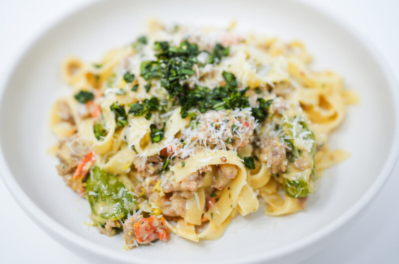 A plate of Creamy Italian Sausage and Shishito Pepper Tagliatelle, showcasing ribbons of pasta coated in a rich, creamy sauce dotted with slices of spicy Italian sausage and tender shishito peppers, garnished with fresh basil and a sprinkle of lemon zest.