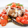 A beautifully plated Mediterranean Lamb, Eggplant, and Tomato Stack featuring spiced lamb patties on roasted eggplant slices, topped with vibrant grape tomato confit and drizzled with creamy tahini sauce.
