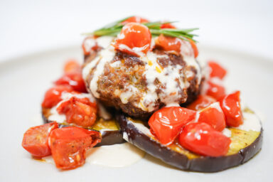 A beautifully plated Mediterranean Lamb, Eggplant, and Tomato Stack featuring spiced lamb patties on roasted eggplant slices, topped with vibrant grape tomato confit and drizzled with creamy tahini sauce.