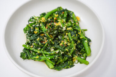 A vibrant dish of spicy garlic rapini, featuring bright green broccoli rabe sautéed with red pepper flakes and garnished with lemon zest, served in a white bowl.