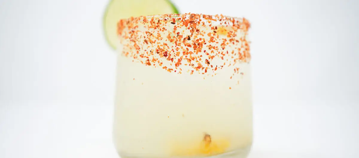 A Sweet Corn Margarita on a clean white background, featuring a glass rimmed with Tajín and topped with roasted corn kernels, highlighted by the vibrant yellow of the corn syrup and a slice of lime for a splash of color.