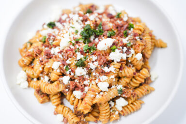 A bowl of Bacon, Mezcal, and Sun-Dried Tomato Rotini, featuring rotini pasta in a creamy mezcal sauce with sun-dried tomatoes, topped with crispy bacon, crumbled cotija cheese, and fresh basil.