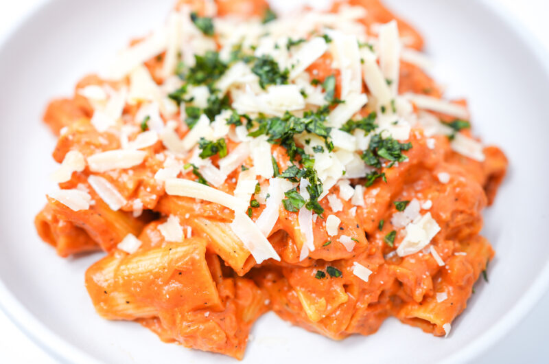 A plate of Garlic Butter Vodka Rigatoni, garnished with freshly grated Parmesan cheese and vibrant green basil leaves. The rigatoni pasta is coated in a rich, creamy sauce, with visible specks of garlic and a hint of red from the pureed tomatoes.