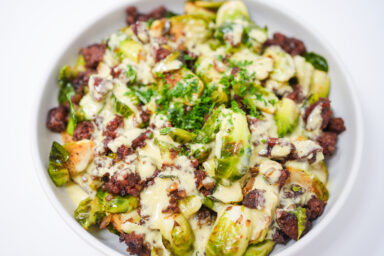 A vibrant dish featuring golden-brown caramelized Brussels sprouts and spicy chorizo slices, topped with a creamy, pale yellow garlic aioli, and garnished with freshly chopped parsley in a white bowl.