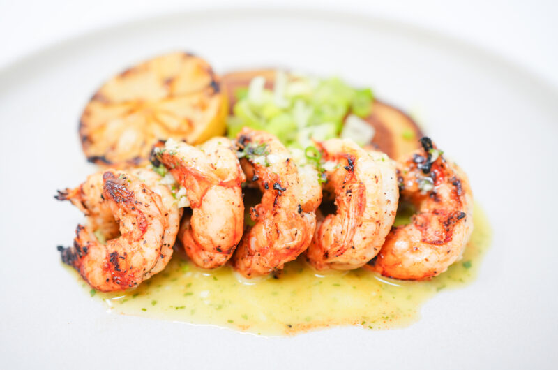 A plate of Citrus Grilled Shrimp skewers, served with golden cornmeal pancakes, a grilled lemon half, and drizzled with creamy cilantro-lime aioli.