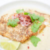 A beautifully plated dish of Pan-Seared Corvina with Coconut Lime Sauce, featuring golden-brown fish fillets atop a creamy, tangy sauce, garnished with vibrant pickled onions, fresh cilantro, lime wedges, and a sprinkle of red pepper flakes.
