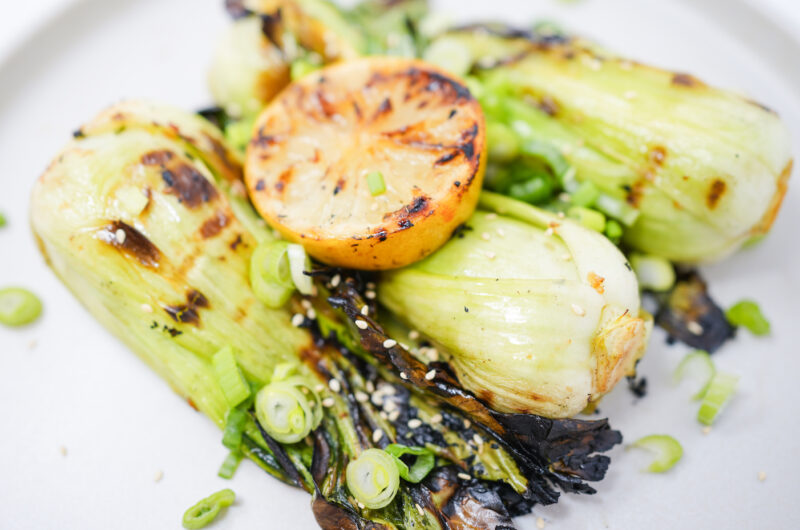 A plate of Garlic Soy Grilled Bok Choy, beautifully charred and topped with grilled lemon halves, sliced green onions, and toasted sesame seeds.