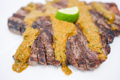 A perfectly grilled porterhouse steak topped with a vibrant, jerk-inspired chimichurri sauce made from fresh herbs, garlic, hot peppers, and warm spices, served on a white plate.