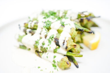 A plate of Garlic Lemon Grilled Okra drizzled with creamy tahini dressing, garnished with fresh parsley and lemon zest. The okra is slightly charred and arranged on a white plate, showcasing its vibrant green color and the smooth, rich tahini sauce.