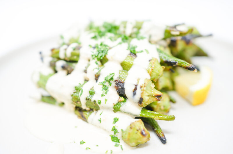 A plate of Garlic Lemon Grilled Okra drizzled with creamy tahini dressing, garnished with fresh parsley and lemon zest. The okra is slightly charred and arranged on a white plate, showcasing its vibrant green color and the smooth, rich tahini sauce.