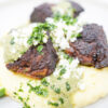 A plate of Pork Carnitas with Green Mole and Mashed Potatoes, featuring tender, crispy pork pieces on a bed of creamy mashed potatoes, drizzled with vibrant green mole sauce, and topped with crumbled queso fresco and fresh cilantro, garnished with lime wedges.