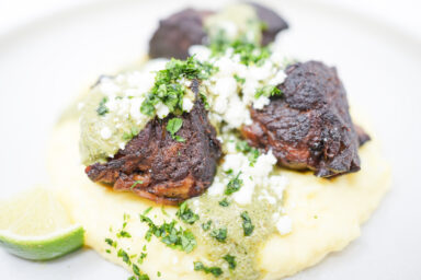 A plate of Pork Carnitas with Green Mole and Mashed Potatoes, featuring tender, crispy pork pieces on a bed of creamy mashed potatoes, drizzled with vibrant green mole sauce, and topped with crumbled queso fresco and fresh cilantro, garnished with lime wedges.