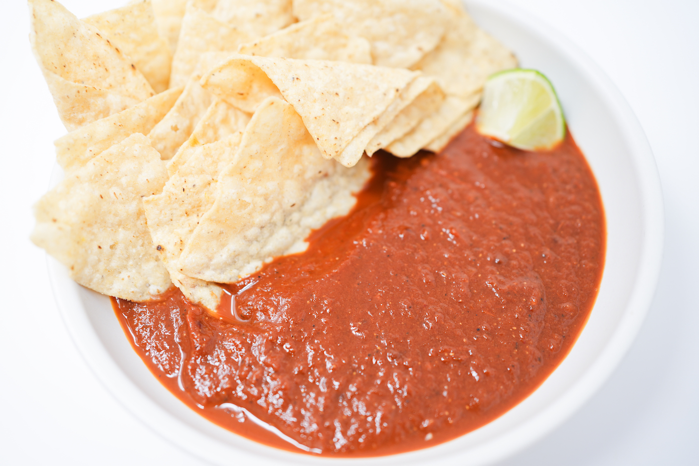 A bowl of vibrant roasted tomato and pasilla pepper salsa, surrounded by crispy tortilla chips and a lime wedge on the side.