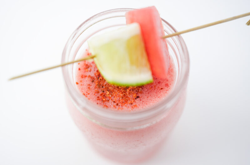 A glass of Watermelon Mezcal Cooler, a vibrant cocktail garnished with a Tajin-rimmed glass, a lime wedge, and a watermelon cube, showcasing a refreshing mix of mezcal, Aperol, lime, and watermelon.