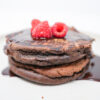 A stack of Chocolate Chipotle Olive Oil Pancakes drizzled with rich chocolate syrup, garnished with fresh raspberries and a sprinkle of ground chipotle pepper.