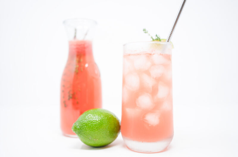 A refreshing glass of Watermelon Thyme Agua Fresca, garnished with a sprig of thyme and a lime wedge, with a vibrant pink hue and served over ice cubes, creating a perfect summer drink